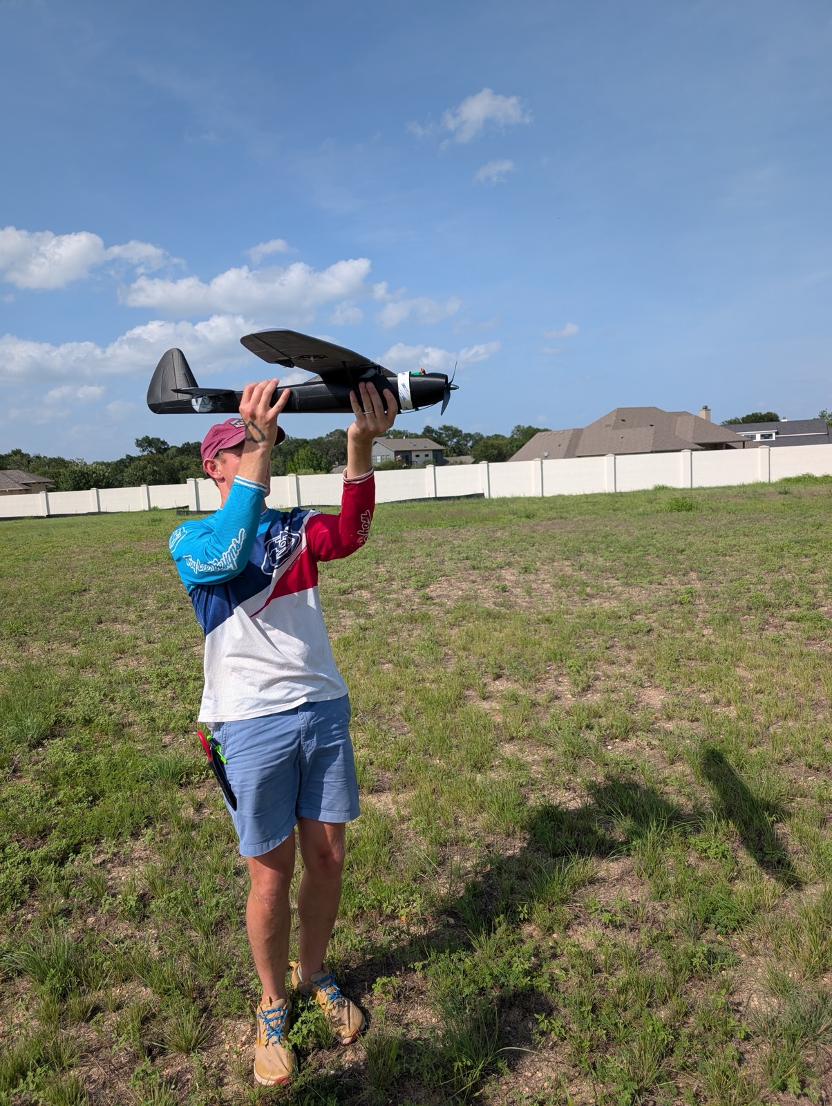 A member in a red cap holding a dark foam plane overhead in a sunny field