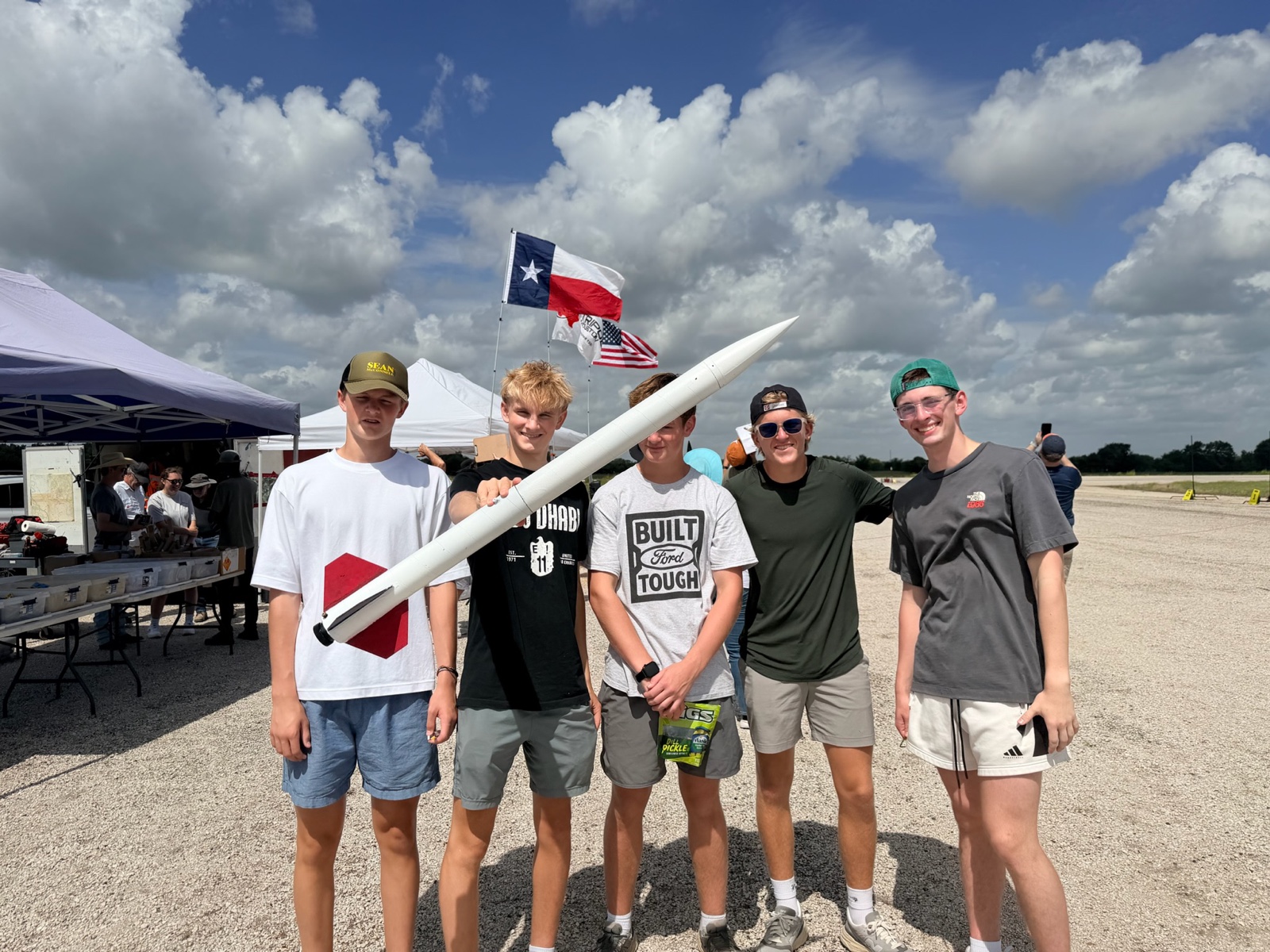 Five club members standing in a row at the launch field, one holding the white rocket at an angle, with Texas and U.S. flags behind them